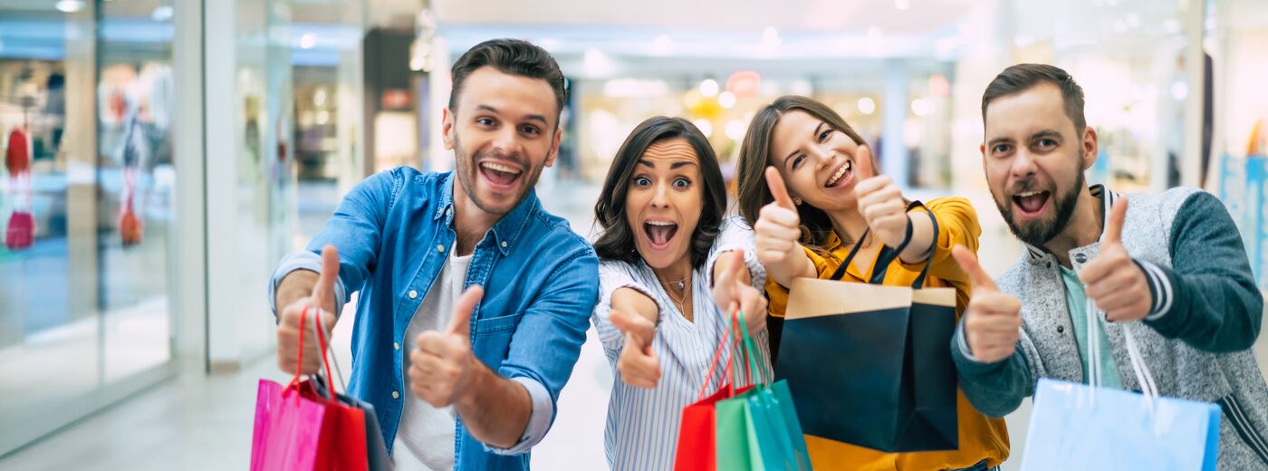 Happy company of smiling excited beautiful stylish friends with colorful paper bags are having fun and showing thumbs up on camera while shopping in the mall.