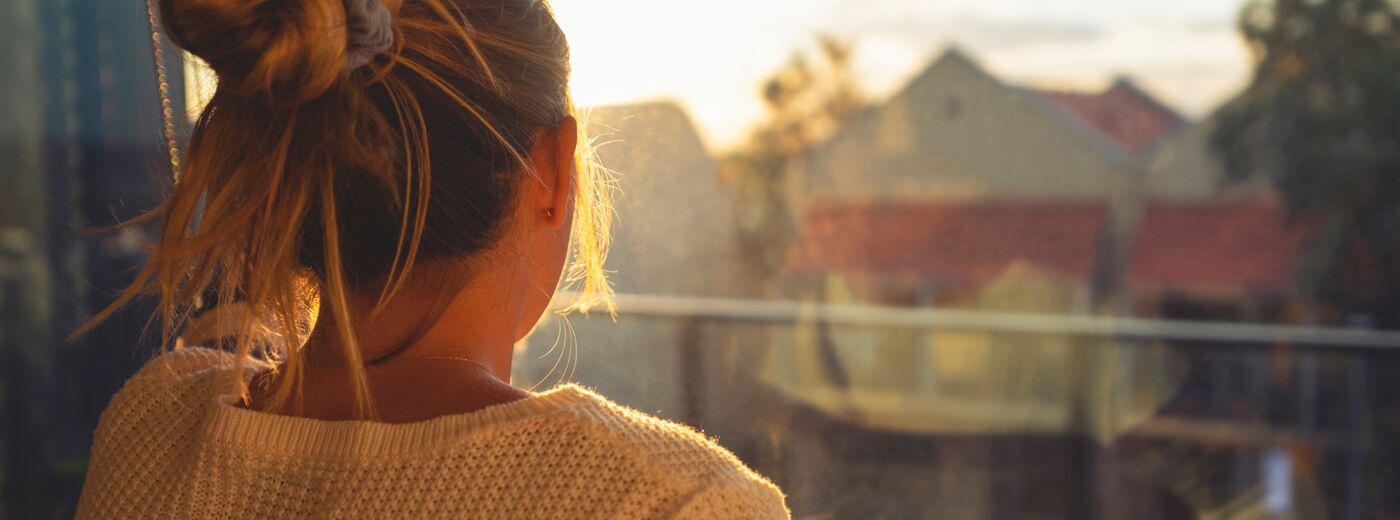 Woman looking through the window at sunset. She is alone and looks a little ad or depressed.