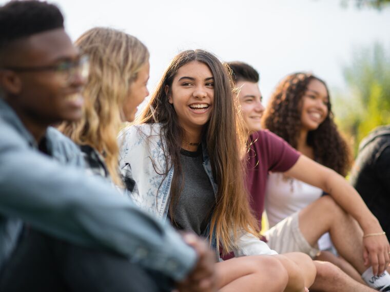 A group of multi-ethnic college and university students sit outside casually hanging out together while on a break.  They are sitting in the grass and laughing together.  Two girls on the far left are looking directly at one another and laughing, and a peer on the far right is leaning over to look at them.  The other male students in the photo are just casually looking straight ahead across the field.