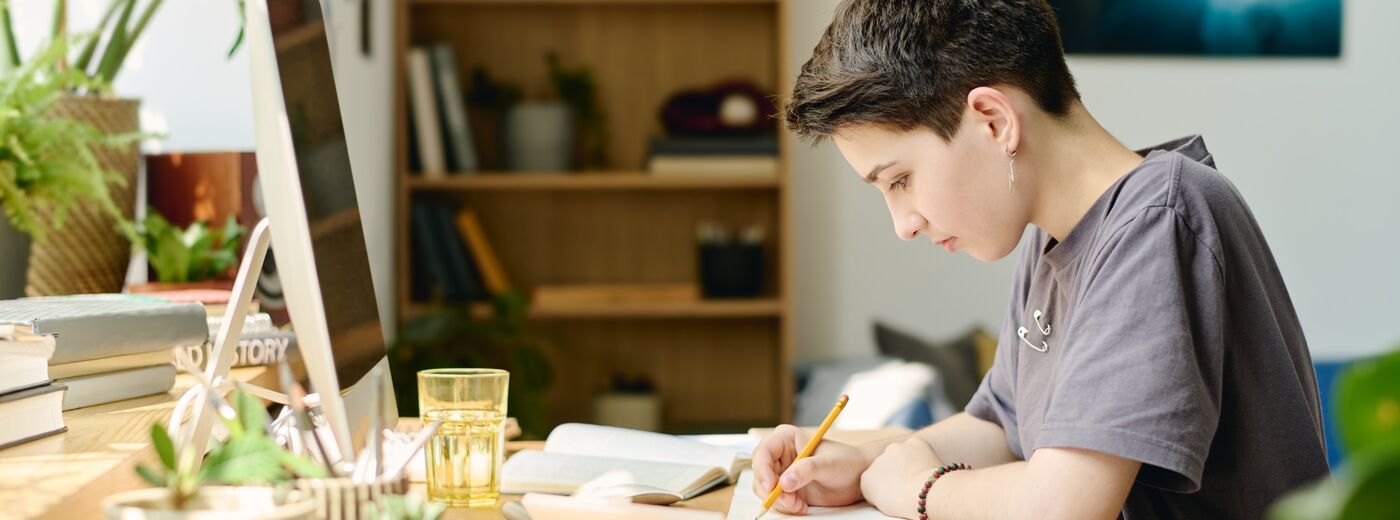 Side view of brunette teenage girl with pencil drawing sketch or making notes on paper while sitting by desk in her room and doing homework