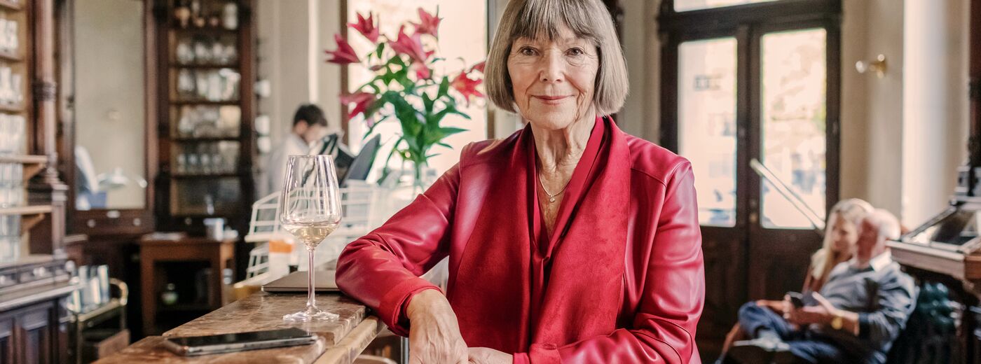 A portrait of a mature woman wearing a red leather jacket sitting at a restaurant bar drinking wine.