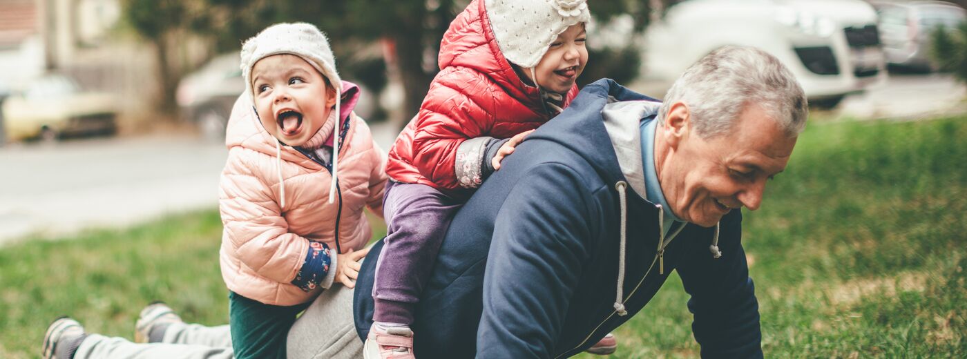 A photo of a playful grandfather and granddaughter. They are casually dressed and playing in the park. They exercise together. A grandfather is exercising while granddaughters are sitting on his back.