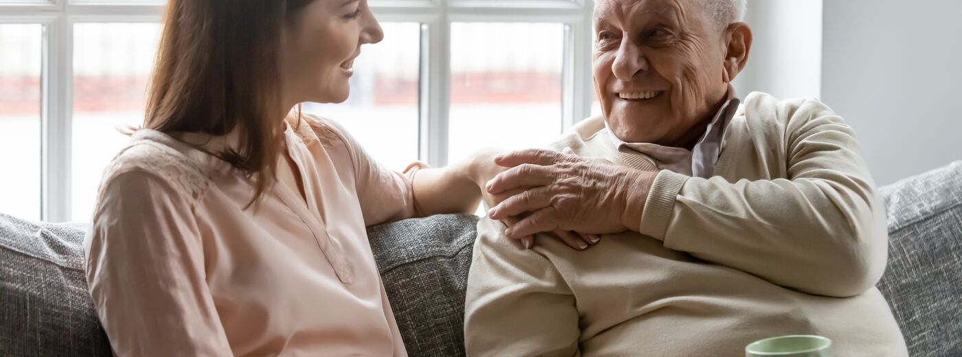 Smiling young woman sitting on sofa with happy older retired 70s father, enjoying pleasant conversation with cup of coffee tea together in living room, mature parents and grown children communication.