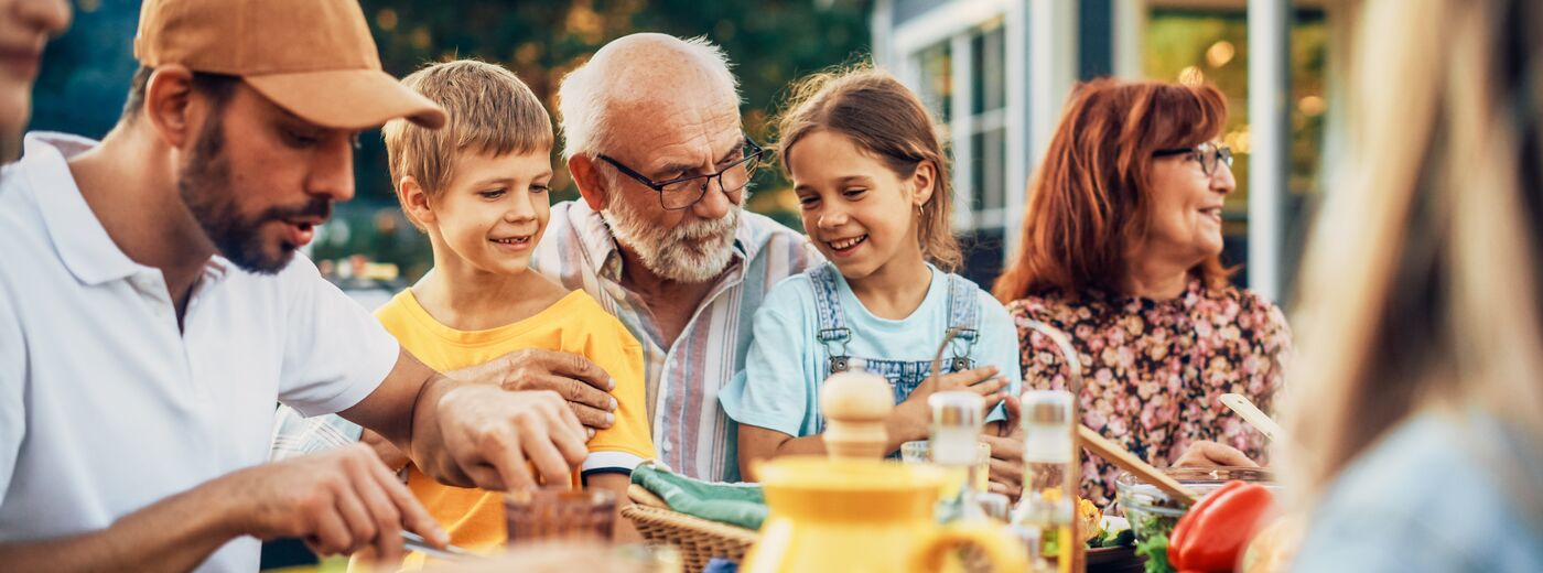 Portrait of a Happy Senior Grandfather Holding His Bright Talented Little Grandchildren on Lap at a Outdoors Dinner Party with Food and Drinks. Family Having a Picnic Together with Children.